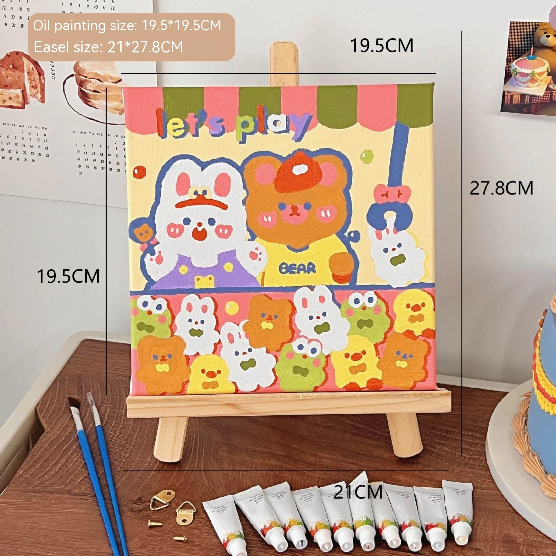 Mini DIY canvas painting kit featuring colourful “Let’s Play” bear design on a small wooden easel, with acrylic paint tubes and brushes arranged on a desk ready for painting