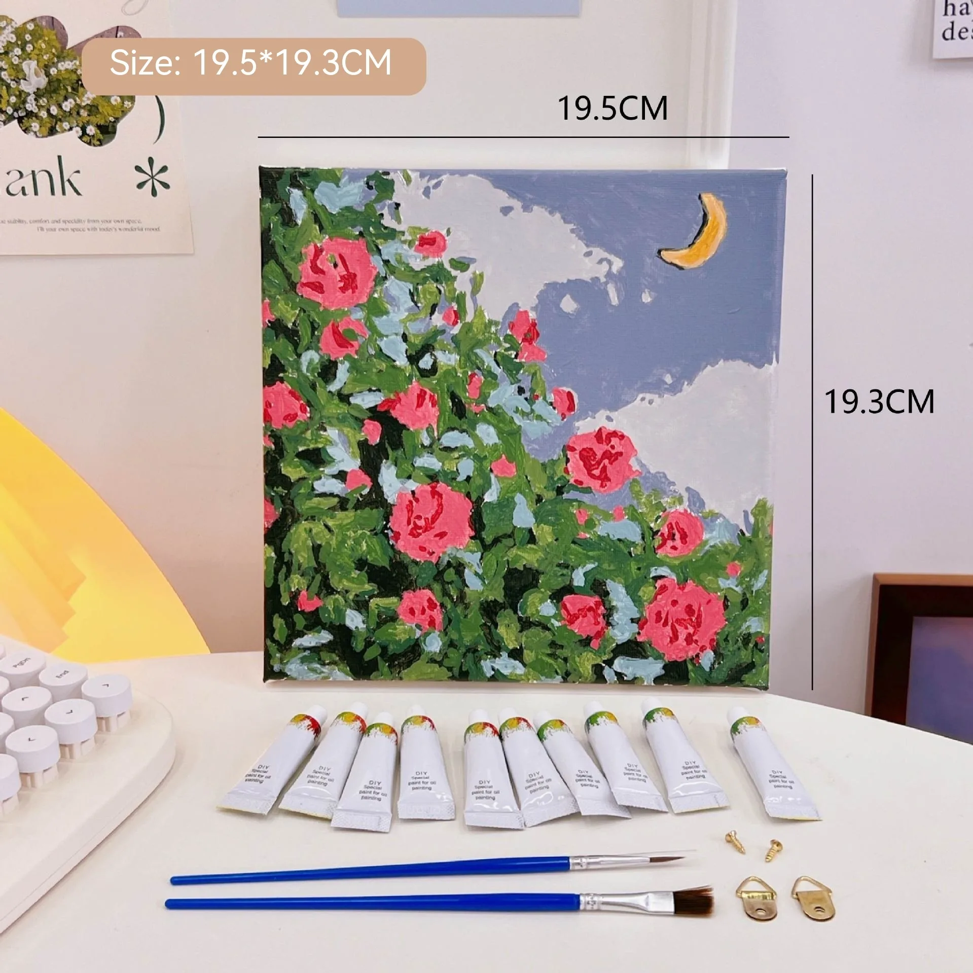 Mini DIY canvas painting kit with pink rose garden and crescent moon design on a pre-printed canvas, including acrylic paint tubes and brushes arranged on a desk ready for painting