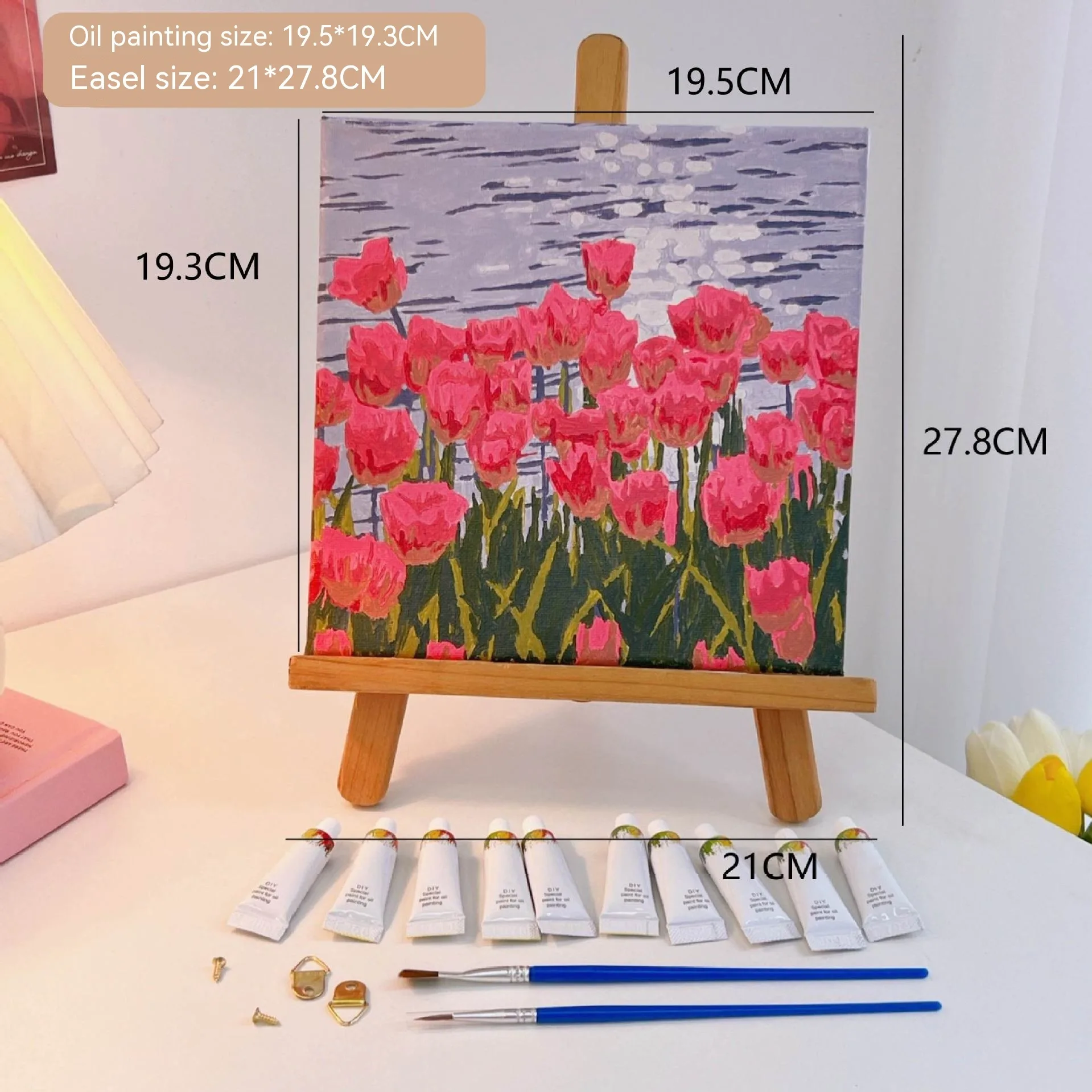 Mini DIY canvas painting kit featuring pink tulips by rippling water on a small wooden easel, with acrylic paint tubes and brushes arranged on a desk ready for painting