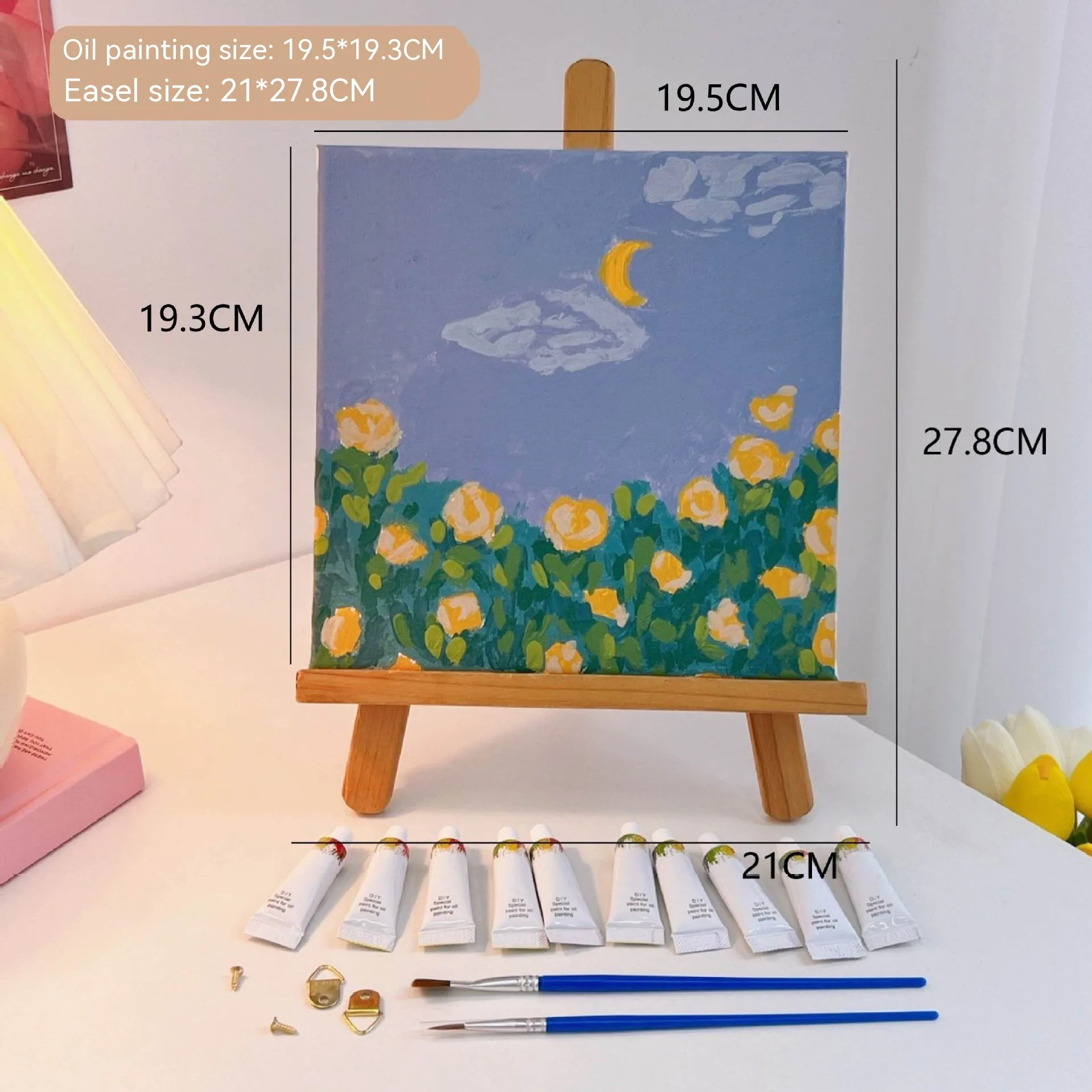 Mini DIY canvas painting kit featuring yellow rose garden under a crescent moon on a small wooden easel, with acrylic paint tubes and brushes arranged on a desk ready for painting