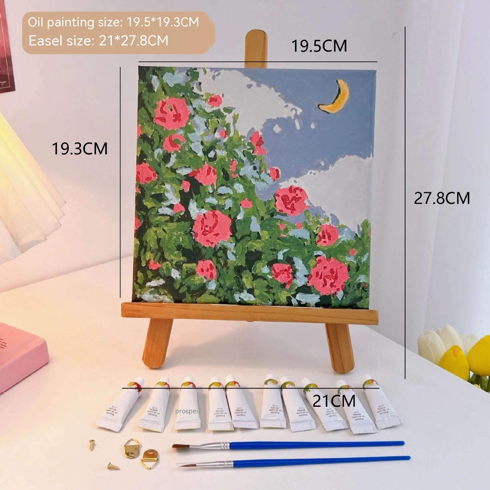 Mini DIY canvas painting kit featuring pink rose garden artwork with a crescent moon on a small wooden easel, with acrylic paint tubes and brushes arranged on a desk ready for painting