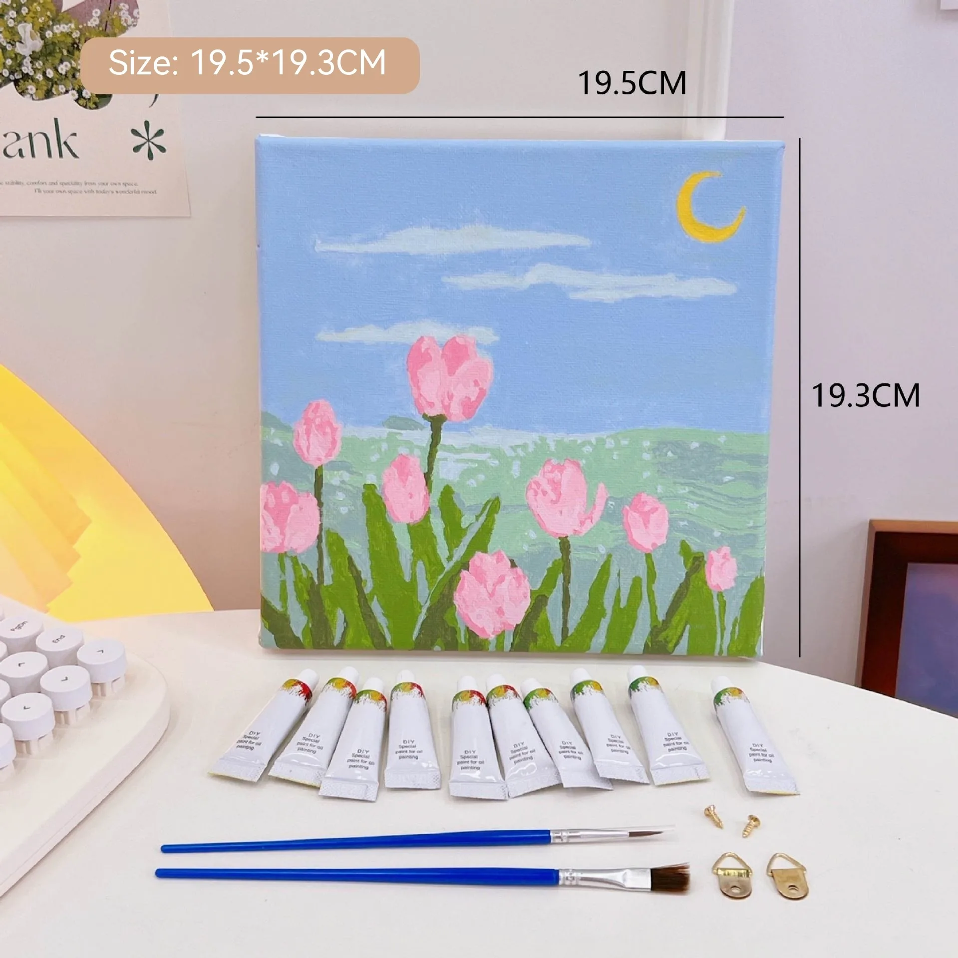 Mini DIY canvas painting kit with pink tulips by the sea under a crescent moon on a pre-printed canvas, including acrylic paint tubes and brushes arranged on a desk ready for painting