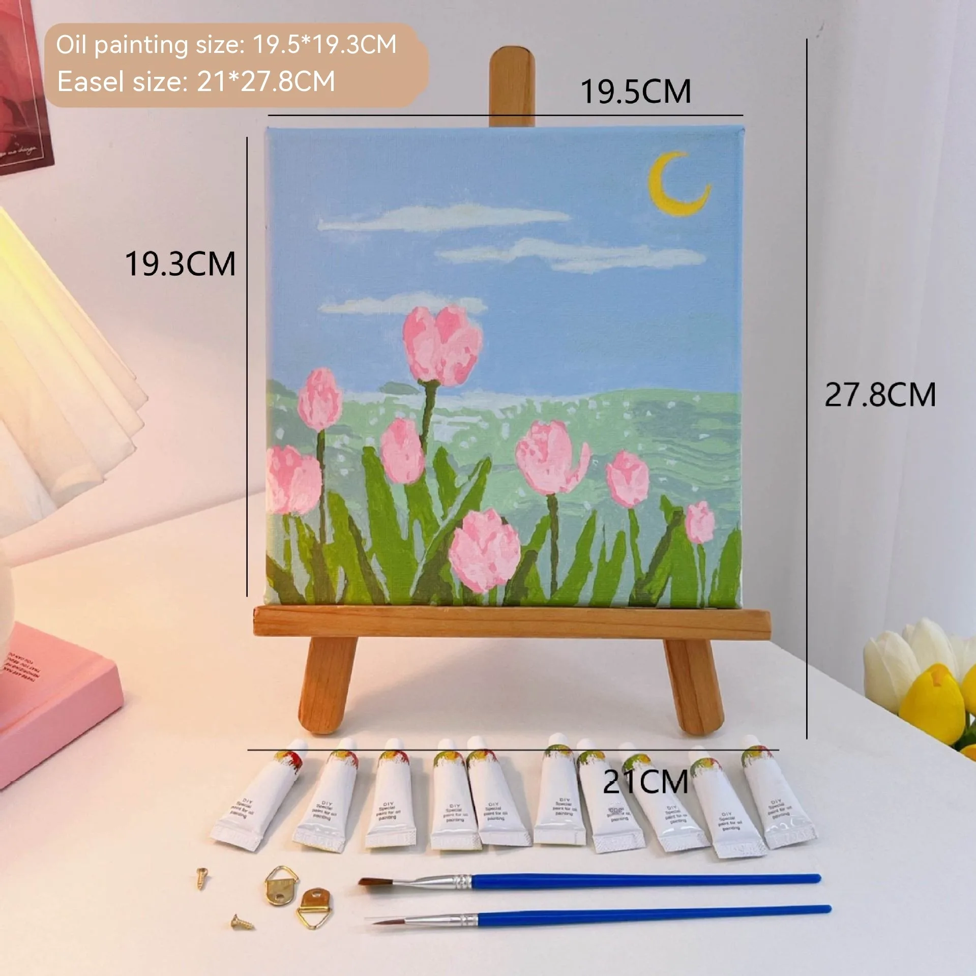 Mini DIY canvas painting kit featuring pink tulips by the sea under a crescent moon on a small wooden easel, with acrylic paint tubes and brushes arranged on a desk ready for painting