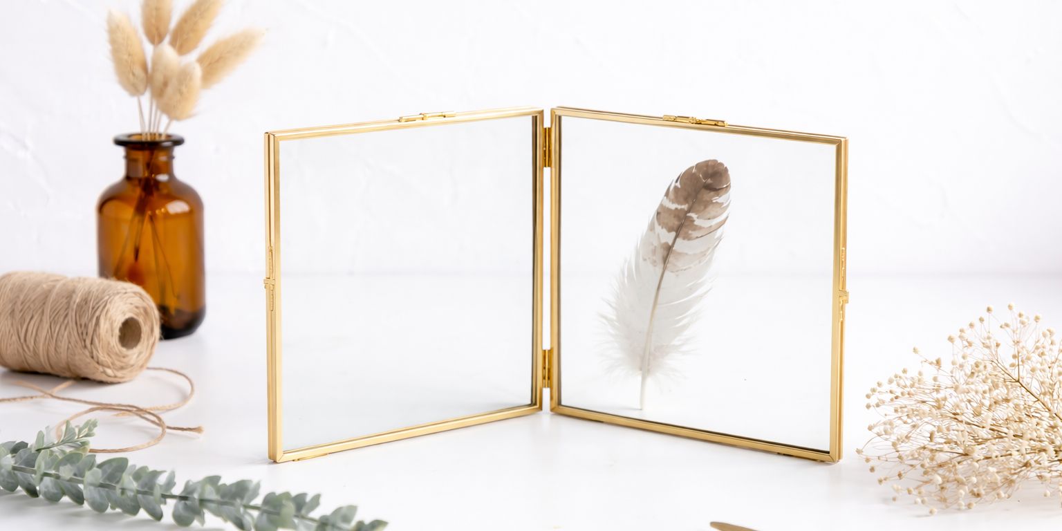 Copper glass folding frame with a feather displayed between double glass panels on a white tabletop with natural décor in a horizontal hero image