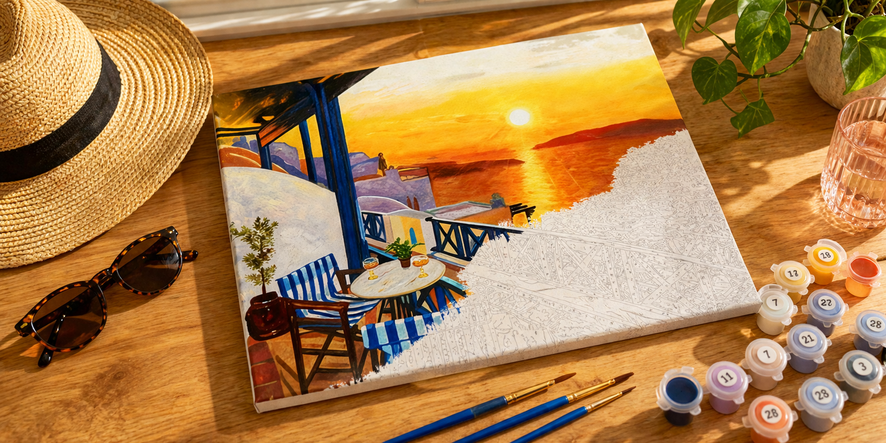 Half-completed sunset paint by numbers canvas on a wooden desk with paint pots, brushes, sunglasses and a straw hat in warm natural sunlight