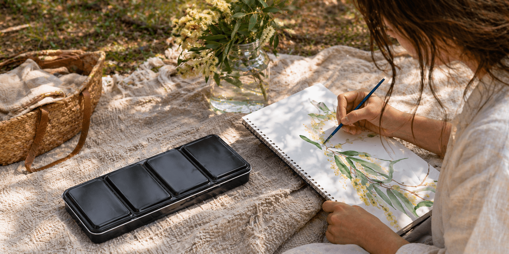 Watercolour tin set in use outdoors on a picnic blanket with artist painting in a sketchbook in natural sunlight